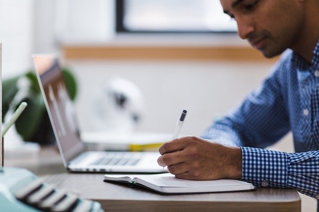 man taking notes on paper from websites on his laptop